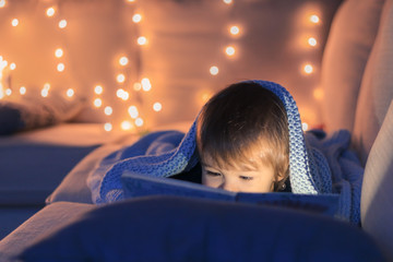 Cute little baby boy reading a book lying on sofa under knitted blanket with garland lights at...