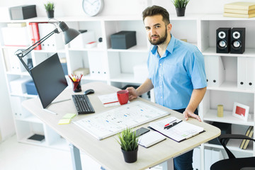 Obraz premium Młody mężczyzna siedzi w biurze przy stole komputerowym, pije kawę i pracuje z tablicą magnetyczną.