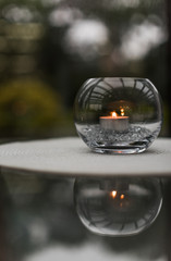 A decorative glass bowl with a candle and filler inside, standing on a white woven mat and reflected from the glass table, with greenery in background, lit with soft evening light, selective focus.