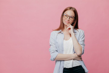 Indoor shot of thoughtful Caucasian woman keeps one hand under chin, dressed in elegant shirt, models over pink background, isolated over pink background with copy space for your promotion or slogan