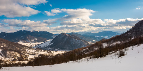 Fototapeta na wymiar panorama of winter countryside in mountains. wonderful sunny day with gorgeous cloudscape. dark forest on the snow covered hills. village down in the distant valley. faraway ridge with snowy tops