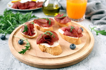 Fototapeta na wymiar Canape or crostini with toasted baguette, light cheese, bresaola and arugula on wooden board. Diet food. Iron food.
