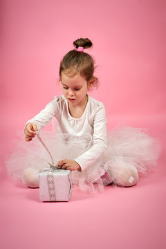 Cute little girl in white tulle skirt opens a gift on a pink background