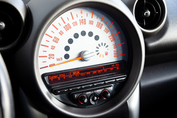 Sensor with a display closeup on the dashboard of a black color on-board computer speedometer with a red arrow and orange numbers and symbols on a white background measuring the vehicle speed.