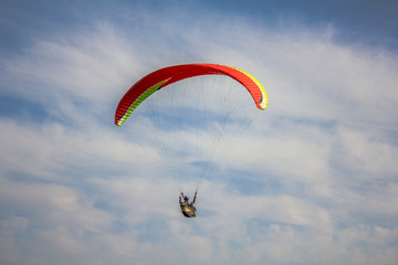 a paraglider on a red with yellow stripes paraglider flies in a blue sky with white clouds