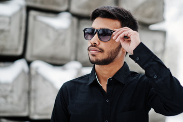 Casual young indian man in black shirt and sunglasses posed against stone blocks.