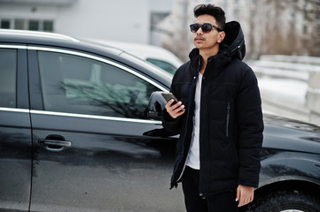Casual young indian man in black jacket and sunglasses posed against suv car with phone at hands.