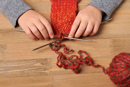 Child girl is learning to knit a scarf. Red wool yarn is on the wooden table. Hand closeup.