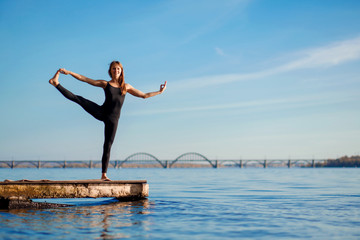 Fototapeta premium Young woman practicing yoga exercise at quiet wooden pier with city background. Sport and recreation in city rush