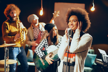 Naklejka premium Mixed race woman singing. In background band playing instruments. Home studio interior.