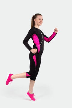 Studio shot young attractive  women in black hundreds of tracksuit with pink accents against  a gray background.  She cheerfully runs with  headphones listening to music and smils
