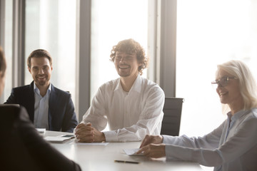 Positive millennial well-dressed businessmen middle aged attractive businesswoman sitting around the office table during negotiations or conference having pleasant conversation with business partners