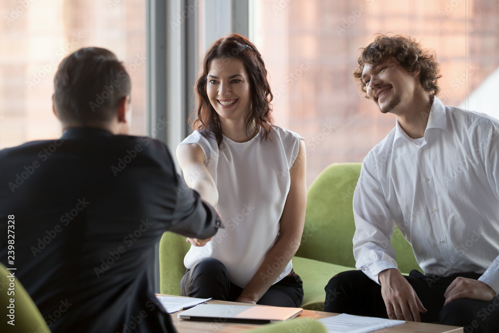 Poster Young professionals company members greeting customer gathered together at business meeting sitting on comfortable couch in modern office. Female employee handshaking with investor expressing respect