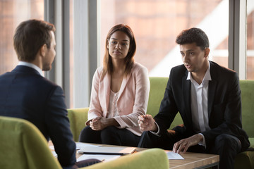 Serious biracial businesswoman and middle eastern ethnicity business partners listening caucasian businessman sitting in comfortable couches negotiating discussing startup new project in modern office