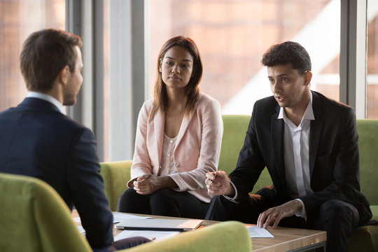 Serious biracial businesswoman and middle eastern ethnicity business partners listening caucasian businessman sitting in comfortable couches negotiating discussing startup new project in modern office