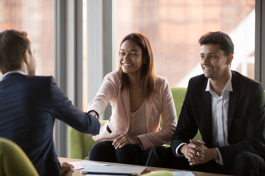 Positive millennial middle eastern ethnicity businessman and african american businesswoman greeting company client, customer rear view, sitting on couches shaking hands ready to start conversation