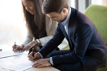Fototapeta premium Positive asian businesswoman sitting on couch with business partner, focus on caucasian businessman, people signing contract agreement document. Successful negotiations, trust and partnership concept