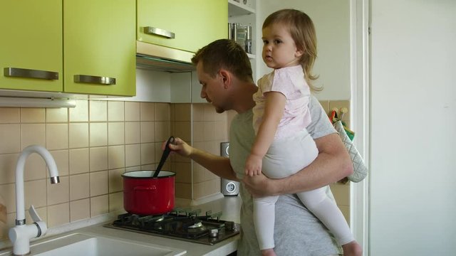 Father's day. Cooking father with small daughter in hands praising son for writing in kitchen, family smiling. Super dad taking care of toddler girl and making a meal lauds boy for correct work sheet