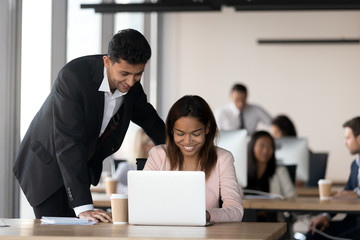 Cheerful multiracial coworkers, middle eastern ethnicity male and african black female looking at computer screen working together in contemporary coworking space with other millennial business people