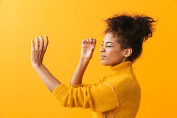 Portrait of beautiful african american woman with afro hairstyle looking through invisible spyglass, isolated over yellow background - obrazy, fototapety, plakaty