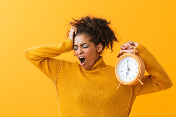 African american unhappy woman in casual clothing yawning while holding alarm clock, isolated over...