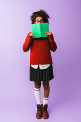 African american young woman in school uniform holding and reading book, isolated over violet background