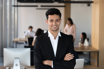 Head shot of confident millennial middle eastern ethnicity office employee or team leader wearing formal suit standing with arms crossed on chest posing looking at camera with coworkers on background.