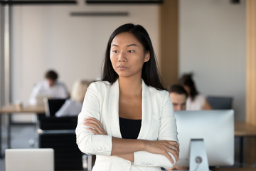 Head shot of serious asian woman stands in coworking office with hands crossed feels dissatisfaction. Pensive businesswoman company employee having difficulties at work thinking about problem solving - obrazy, fototapety, plakaty