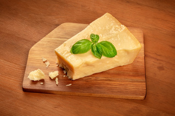A piece of aged Parmesan cheese with crumbs and fresh basil on a dark rustic wooden background with copy space