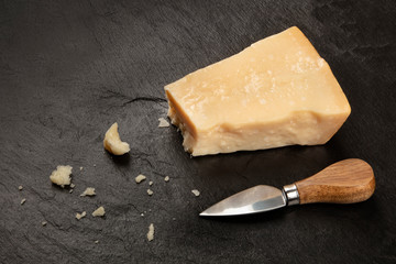A piece of aged Parmesan cheese with crumbs on a black background with a cheese knife and a place for text