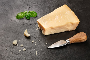 A piece of aged Parmesan cheese with crumbs on a black background with fresh basil leaves, a cheese knife, and copy space