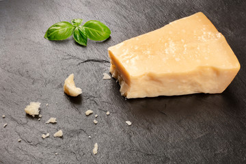 A piece of aged Parmesan cheese with crumbs on a black background with fresh basil leaves and a place for text