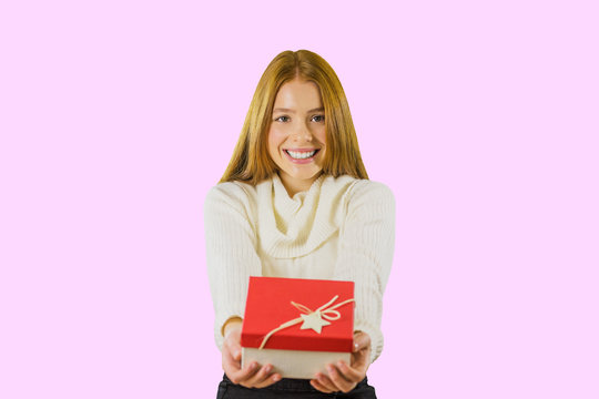 Attractive girl with long red hair look into the camera and holds gift on an isolated pink background