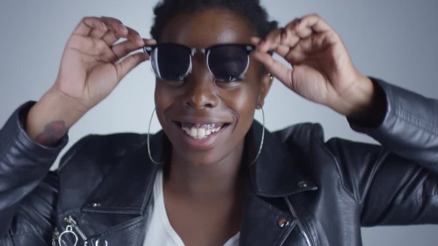 Portrait shot of happy black woman wearing sunglasses, hoops earrings and leather jacket smiling for camera and dancing in studio with grey background