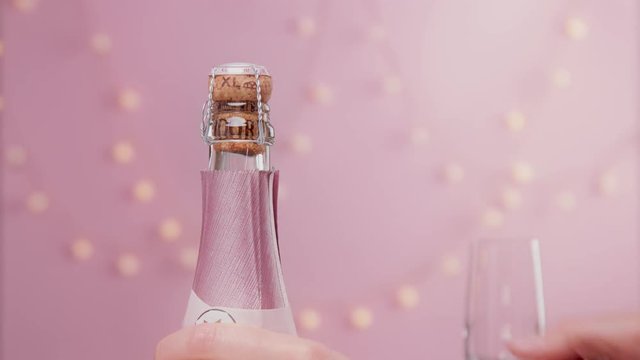 Pink sparkling wine with womans hand and glasses on pink background with christmas lights female christmas celebration closeup of woman hand take of pink foil from champagne bottle