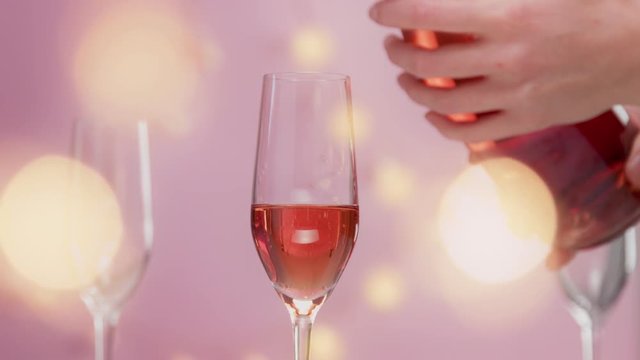 Pink sparkling wine with womans hand and glasses on pink background with christmas lightsslow motion woman hand full glass with rose sparkling wine and lights behind the camera.