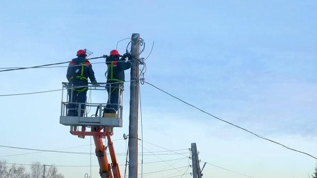 Electricians in uniform repair power lines, standing on the bucket. Winter day. Cold weather. In the background is a blue cloudy sky.