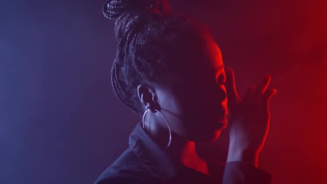 Low-key portrait shot with red and blue lighting of black woman with braids and hoops earrings dancing sensually and touching her face
