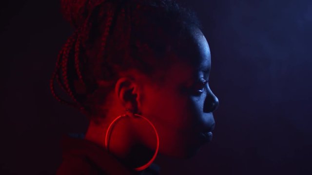 Low-key portrait with red and blue lighting: side view of black woman with braids and hoops earrings standing in dark studio with her head thrown back, then looking forward with determination