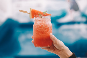 Hand with Fresh watermelon smoothie in the glass jar. Background is blur swimming pool