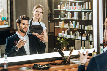 smiling young hairstylist and male client looking at mirror in beauty salon
