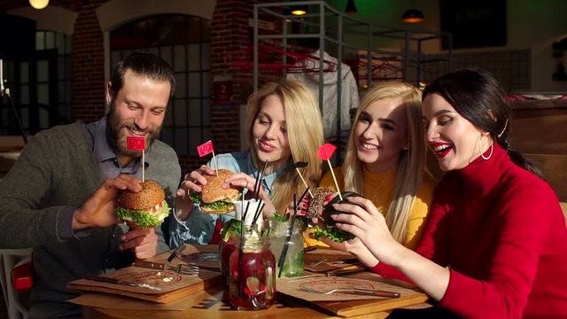 Cheerful young friends eat burgers in the cafe. Four teenagers are holding large juicy hamburgers. A group of people eating fast food in a modern restaurant. Slow motion.