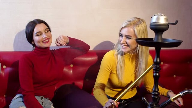 Two sexy girls smoke hookah in a cafe, they sit on a big red sofa and let out a lot of smoke from the mouth. A group of friends relax in a cafe, they talk, laugh and smoke a hookah. Slow motion.