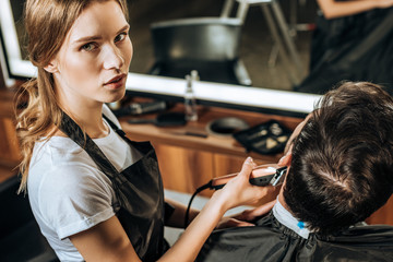 high angle view of young hairdresser using electric hair clipper and looking at camera while working with client in beauty salon - obrazy, fototapety, plakaty