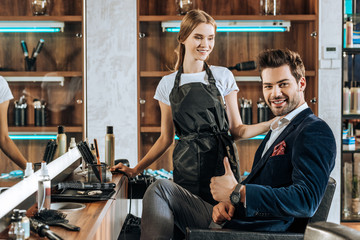 smiling young hairdresser looking at handsome happy client showing thumb up in beauty salon