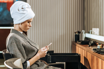 attractive young woman with towel on head sitting and using smartphone in beauty salon