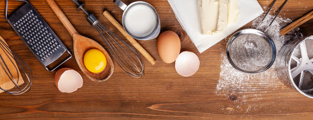 Kitchen, pastry inventory lies on the wooden table. Butter, eggs, wooden spoons, whisk, flour, milk. Top view with copy space, mockup for menu, recipe for culinary classes. Banner