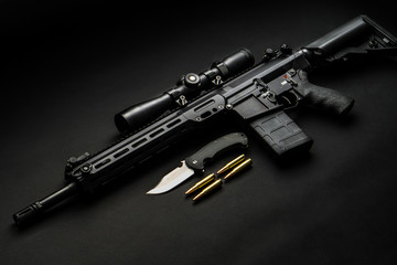 modern automatic rifle with an optical sight lies on a dark background and next to the knife and...