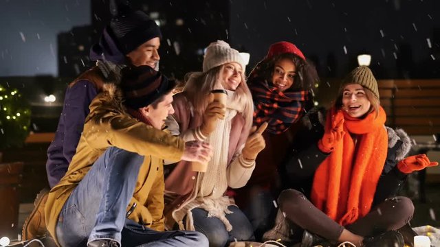 Multiethnic friends in warm clothes drinking coffee and singing in snow street