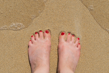 view on female feet with red pedicure on a sunny sandy beach with a wave